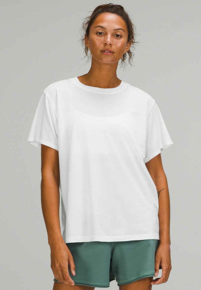 Witte katoenen t-shirt met korte mouwen, ronde hals, relaxte pasvorm, gedragen met groene shorts. Glad textuur, geen zichtbare patronen of accenten.