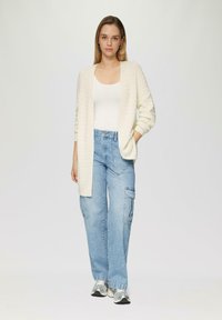 Lange, open-front cardigan in ribbed crèmekleurige brei, gedragen over een fitted witte tanktop. Gecombineerd met lichtblauwe cargobroek en zilveren sneakers.