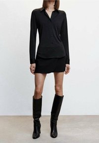 Schwarzes Langarmhemd mit Knopfschluss, kombiniert mit einem schwarzen Minirock und knielangen schwarzen Stiefeln, alles aus einem glatten Stoff.