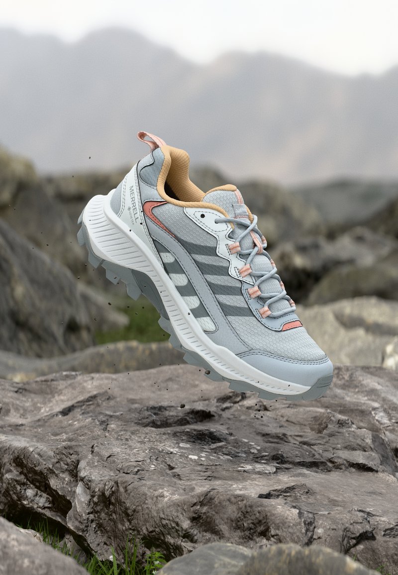 Chaussure de randonnée Merrell grise et pêche avec semelle robuste flottant au-dessus d'un terrain rocheux, avec des montagnes floues en arrière-plan.