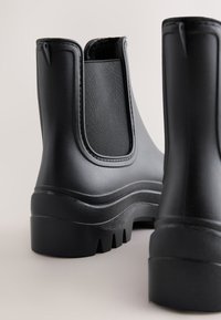 Pair of black rubber ankle boots with elastic side panels and thick treaded soles shown on a neutral background.