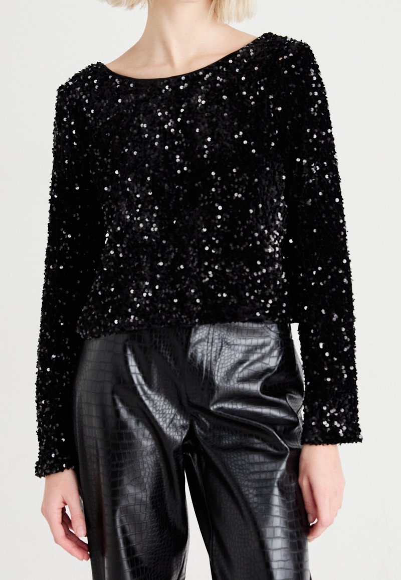 Haut noir à manches longues orné de sequins, associé à un pantalon noir taille haute en simili cuir avec une texture discrète croco.