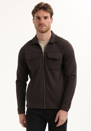 Chaqueta marrón con cremallera hecha de tejido suave, con cuello, dos bolsillos en el pecho y bolsillos laterales. Textura suave y diseño ajustado.