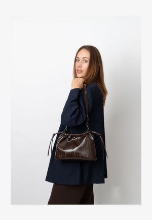 Borsa a mano in pelle marrone lucida con design arricciato, chiusura a coulisse e tracolla regolabile, indossata da una modella su sfondo neutro.