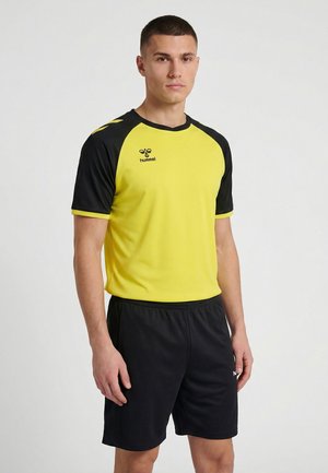 Jonge man die een geel-zwart sportshirt met korte mouwen en zwarte sportshorts draagt, staand tegen een effen witte achtergrond.