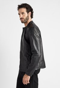 Zwarte leren jas met een rits aan de voorkant, gequiltte schouderdetails, een gladde textuur en een getailleerde pasvorm. Inclusief zijzakken.