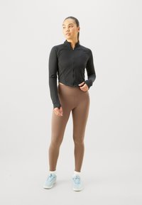 Veste de sport noire courte et leggings marron, avec un design ajusté. Le modèle porte des baskets bleu clair avec des semelles texturées.