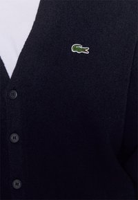 Černý cardigan na knoflíky s malým zeleným výšivkovým logem krokodýla na levé hrudi přes bílou košili.