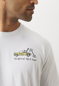 Hvid langærmet t-shirt med et grafisk motiv af en gul bugserbil og teksten "Vi løfter det hele, stort og småt."