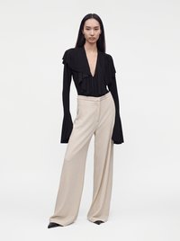 Femme portant une blouse noire à manches longues avec des volants, un décolleté plongeant en V et un pantalon beige à jambes larges, debout devant un fond blanc uni.