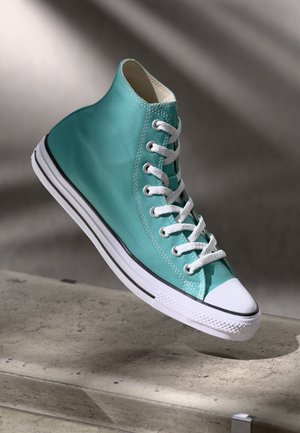 Hoher türkisfarbener Leinensneaker mit weißen Schnürsenkeln und Gummisohle, schwebend über einer Betonfläche mit schattigem Hintergrund.