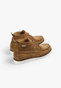 Un par de zapatillas altas informales de ante color beige con suelas blancas, cordones y costuras visibles sobre un fondo blanco.