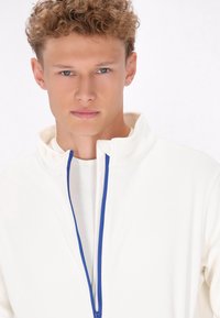 Witte zip-up jas met een hoge kraag, voorzien van een blauwe rits. De stof lijkt glad en heeft een lichte textuur.