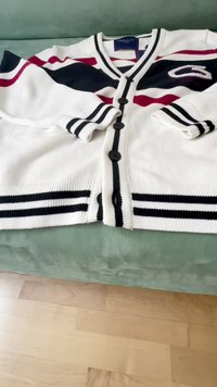 Cárdigan blanco con rayas negras y rojas, textura acanalada, cinco botones negros y dos bolsillos frontales. Etiqueta de la marca visible en el cuello.