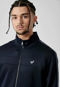 Veste de track zippée marine en tissu doux, avec des bandes latérales blanches et un logo brodé sur la poitrine. Design à col montant.