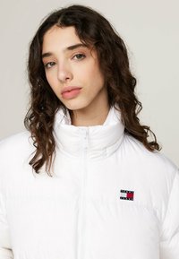 Giacca imbottita bianca con colletto alto, zip frontale e patch logo ricamato con colori rosso, bianco e blu sul petto.