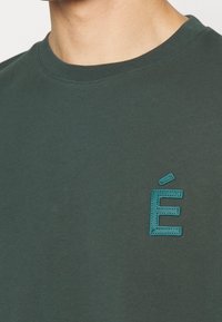 Tmavě zelené bavlněné tričko s texturovanou tyrkysovou výšivkou písmene "É" na levé části hrudi. Krátké rukávy a kulatý výstřih.