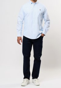 Camisa de color azul claro con botones y cuello, combinada con vaqueros oscuros y zapatillas blancas. Pequeño logo notable en la camisa.