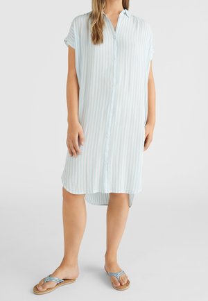 Kobieta ubrana w luźną, do kolan, jasnoniebiesko-białą sukienkę w paski na guziki z krótkimi rękawami oraz jasnoniebieskie sandały, stojąca na białej podłodze.