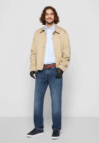 Jachetă bej deschis, cămașă albastră în carouri, blugi albaștri, curea maro, mănuși negre și adidași închiși la culoare. Fundal simplu. Ținută casual.