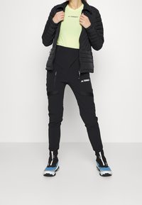 Svart isolerad jacka med dragkedjefickor, grön t-shirt och svarta byxor med remmar och dragkedjefunktioner. Blå och svarta skor med vita detaljer.