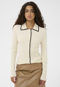 Ženska s kodrastimi lasmi nosi kremast žametan kardigan z črnimi robovi in gumbi, v kombinaciji s krilom iz rjave usnjene tkanine, ki stoji ob beli steni.