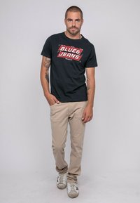 T-shirt en coton noir avec un graphisme "BLUE&JEANS" en rouge et blanc. Un pantalon chino beige et des baskets blanches avec des accents foncés complètent la tenue.