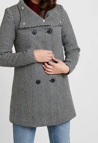Žena v černobílém kabátu s herringbone vzorem a dvojitým zapínáním, s černými knoflíky, drží kabát zavřený a má na sobě modré džíny.