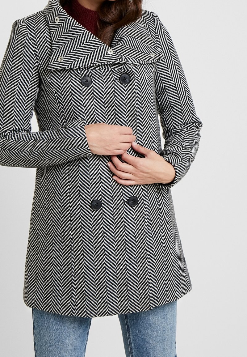 Žena v černobílém kabátu s herringbone vzorem a dvojitým zapínáním, s černými knoflíky, drží kabát zavřený a má na sobě modré džíny.
