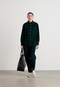 Camicia in corduroy verde scuro con taschino, abbinata a pantaloni coordinati. Il modello porta una borsa tote nera e indossa sneakers argentate.