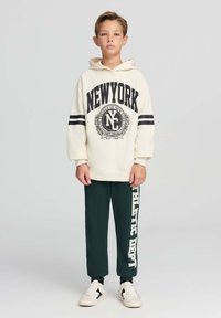 Felpa crema con stampa nera "NEW YORK" e righe, abbinata a pantaloni jogger verde scuro con "ATHLETIC DEPT" in bianco. Sneakers bianche.