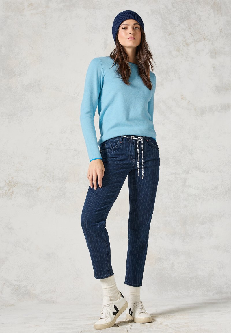 Un maglione azzurro, jeans scuri a righe con un elastico in vita, calzini bianchi e sneakers bianche con dettagli neri. Berretto di lana blu navy.