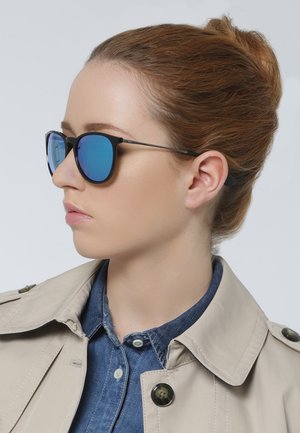 Occhiali da sole neri con lenti blu specchiate e sottili bracci in metallo, indossati da una persona in un trench beige e camicia di denim.