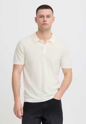 Junger Mann mit kurzen Haaren, der ein weißes Polohemd mit kurzen Ärmeln und schwarze Hosen trägt, steht vor einem schlichten hellen Hintergrund.