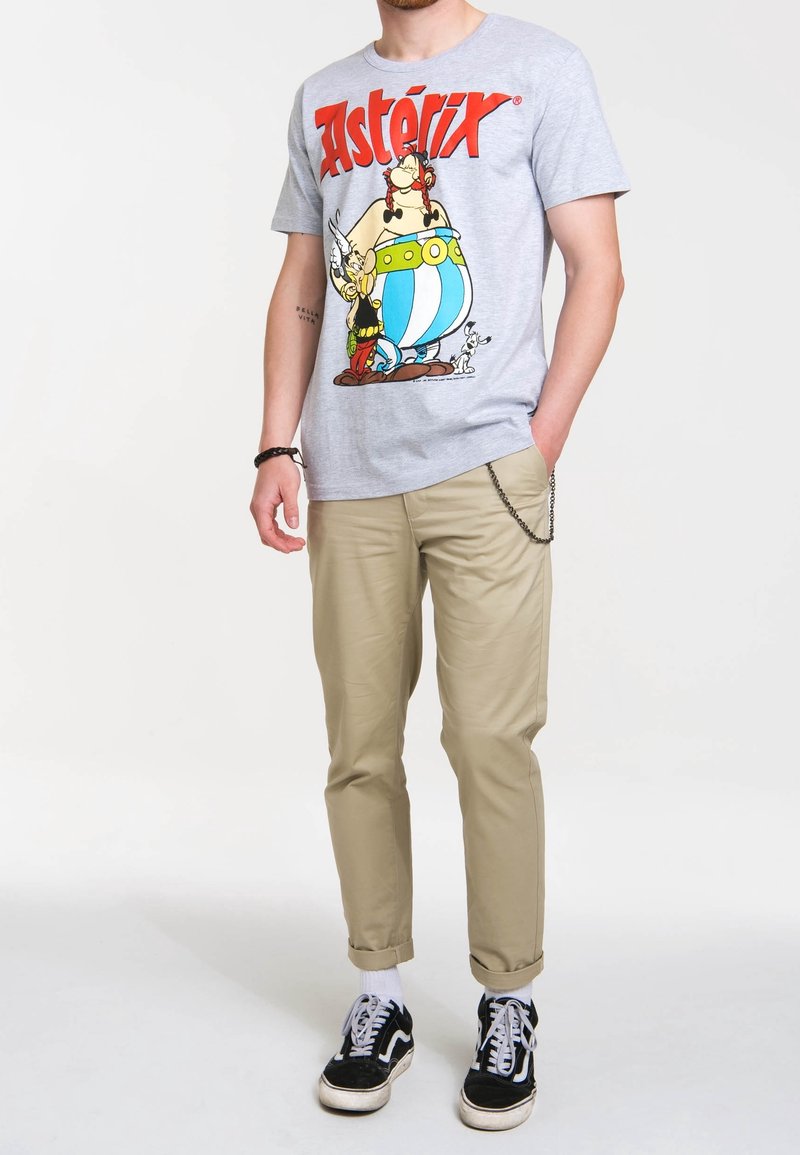 Szary t-shirt z grafiką "Asterix", połączony z beżowymi wąskimi spodniami i czarnymi sneakersami z białymi akcentami.