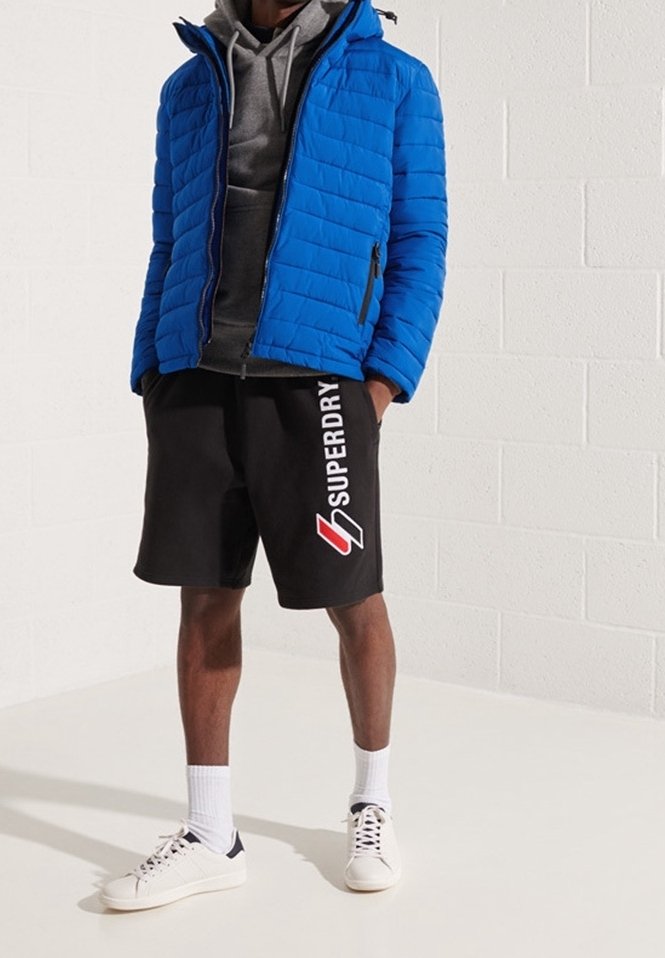 Homme portant une doudoune bleue par-dessus un sweat à capuche gris, un short noir Superdry avec logo, des chaussettes blanches et des baskets blanches, debout contre un mur blanc.