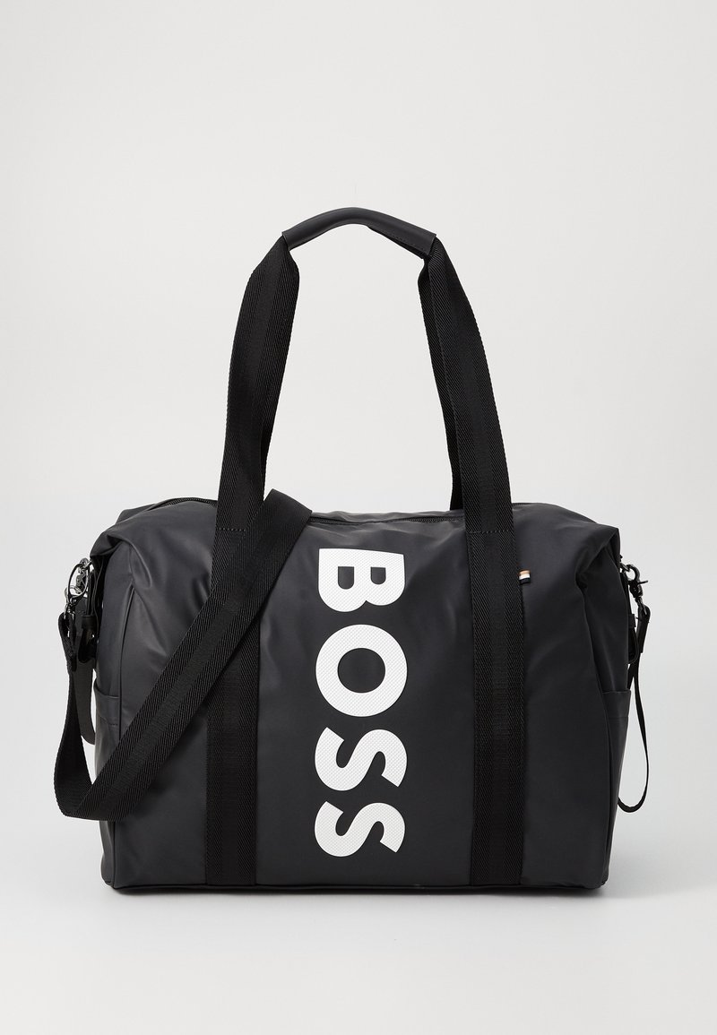 Black duffle bag made of glossy material, featuring large white "BOSS" logo, two top handles, and an adjustable shoulder strap.