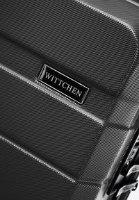 Černý pevný kufr s texturovaným povrchem, vybavený obdélníkovou kovovou štítkem s logem a kombinovaným zámkem na boku.