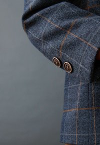 Manche d'un blazer à carreaux bleu avec un motif quadrillé marron et blanc et deux boutons marron sur le poignet sur fond gris.