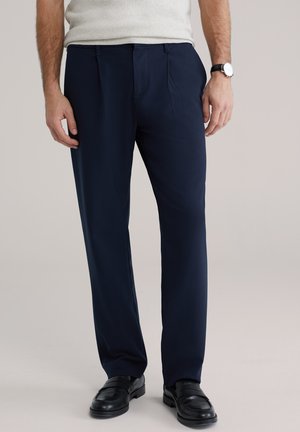 Marineblauwe broek gemaakt van gladde stof, met twee plooien aan de voorkant en een getailleerde pasvorm, gecombineerd met zwarte leren schoenen.