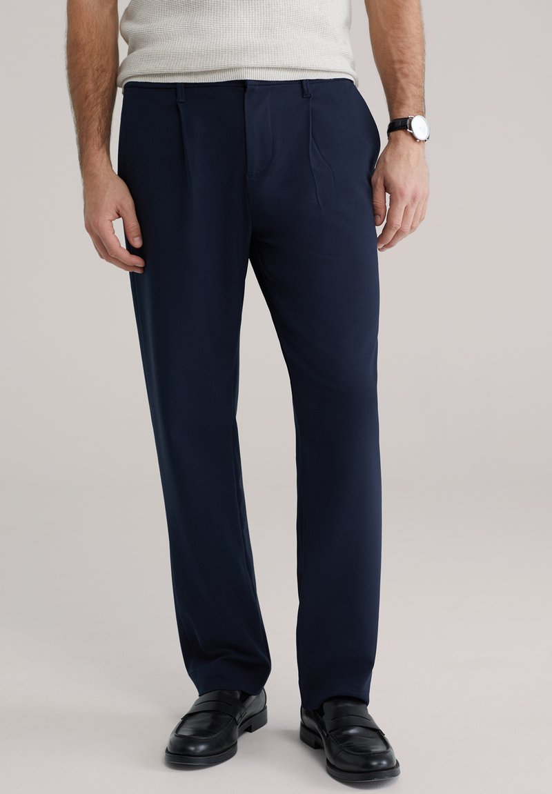 Pantaloni bleumarin din material neted, cu două pliuri frontale și croială ajustată, asortați cu pantofi negri din piele.