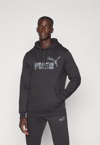 Felpa nera con cappuccio e tasca a marsupio, caratterizzata da un logo Puma riflettente. Vestibilità regolare con tessuto strutturato e pantaloni neri coordinati.