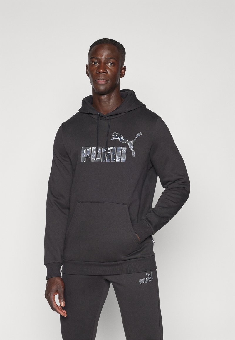 Felpa nera con cappuccio e tasca a marsupio, caratterizzata da un logo Puma riflettente. Vestibilità regolare con tessuto strutturato e pantaloni neri coordinati.