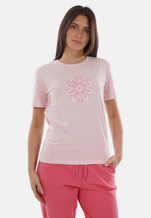 Donna che indossa una maglietta rosa chiaro con un intricato disegno mandala sul petto e pantaloni rosa con coulisse, in piedi davanti a uno sfondo bianco.