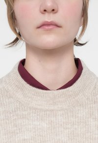 Beige ribbad tröja med hög krage över en vinröd skjorta, som lyfter fram de kontrasterande färgerna och texturerna i tygerna.