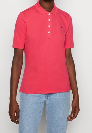 Personne portant un polo rose vif à manches courtes avec quatre boutons et un jean bleu clair, visible du cou aux hanches.