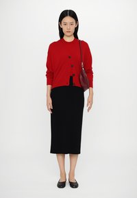Rode cardigan met zwarte knopen over een zwarte midi-rok. Model draagt zwarte flats en heeft een bordeauxkleurige schoudertas.