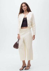 Giacca cropped crema e pantaloni a gamba larga, abbinati a un top corto marrone. Gli accessori includono una borsa marrone. Le calzature sono sandali neri con cinturini.