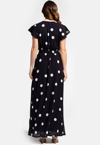 Maxime dress in black fabric with white polka dots, featuring short flutter sleeves, an elastic waist, and a flowing skirt that reaches the ankles.