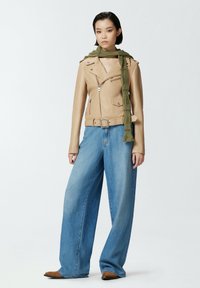 Chaqueta de moto de cuero beige con bolsillos con cremallera, llevada sobre una blusa de cuello en V. Combinada con unos vaqueros de pierna ancha azul y una bufanda de canalé verde.